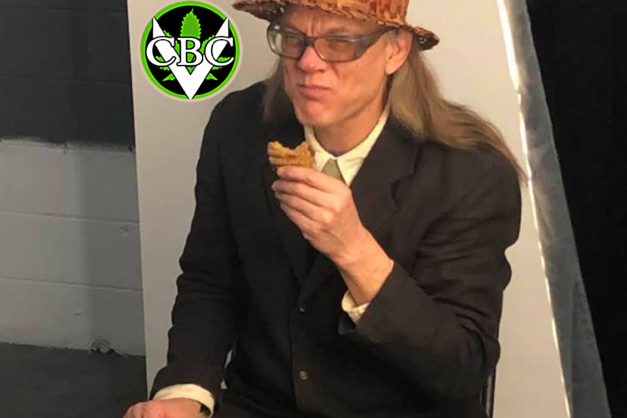 Ted Smith, founder of the Victoria Cannabis Buyers Club, sits on a chair in a photo studio, wearing a woven hat and black suit, smiling slightly while holding a cookie - symbolizing resilience, grassroots leadership, and the enduring spirit of cannabis compassion.